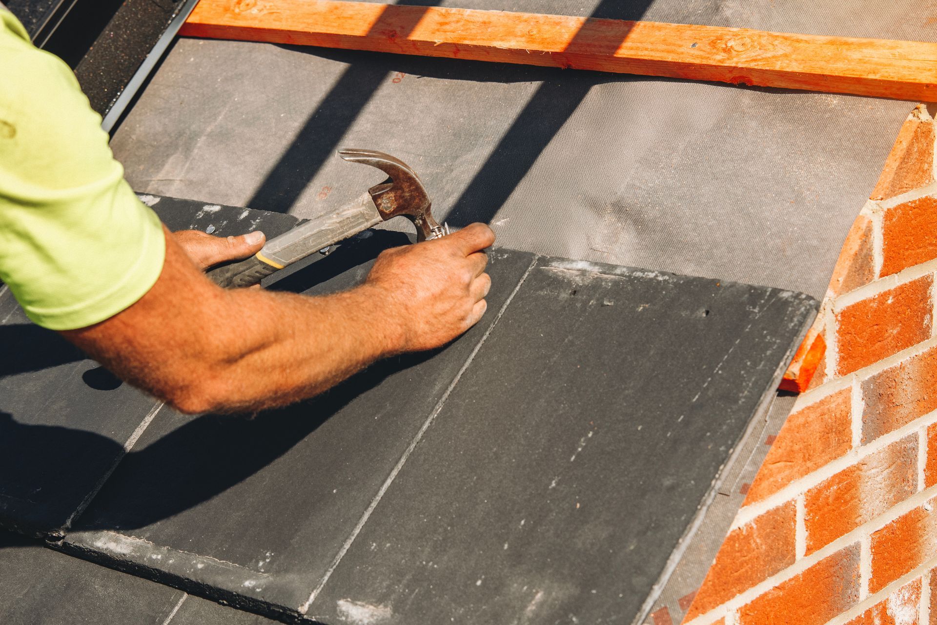 Person hammering roofing shingles on a roof next to an orange brick wall