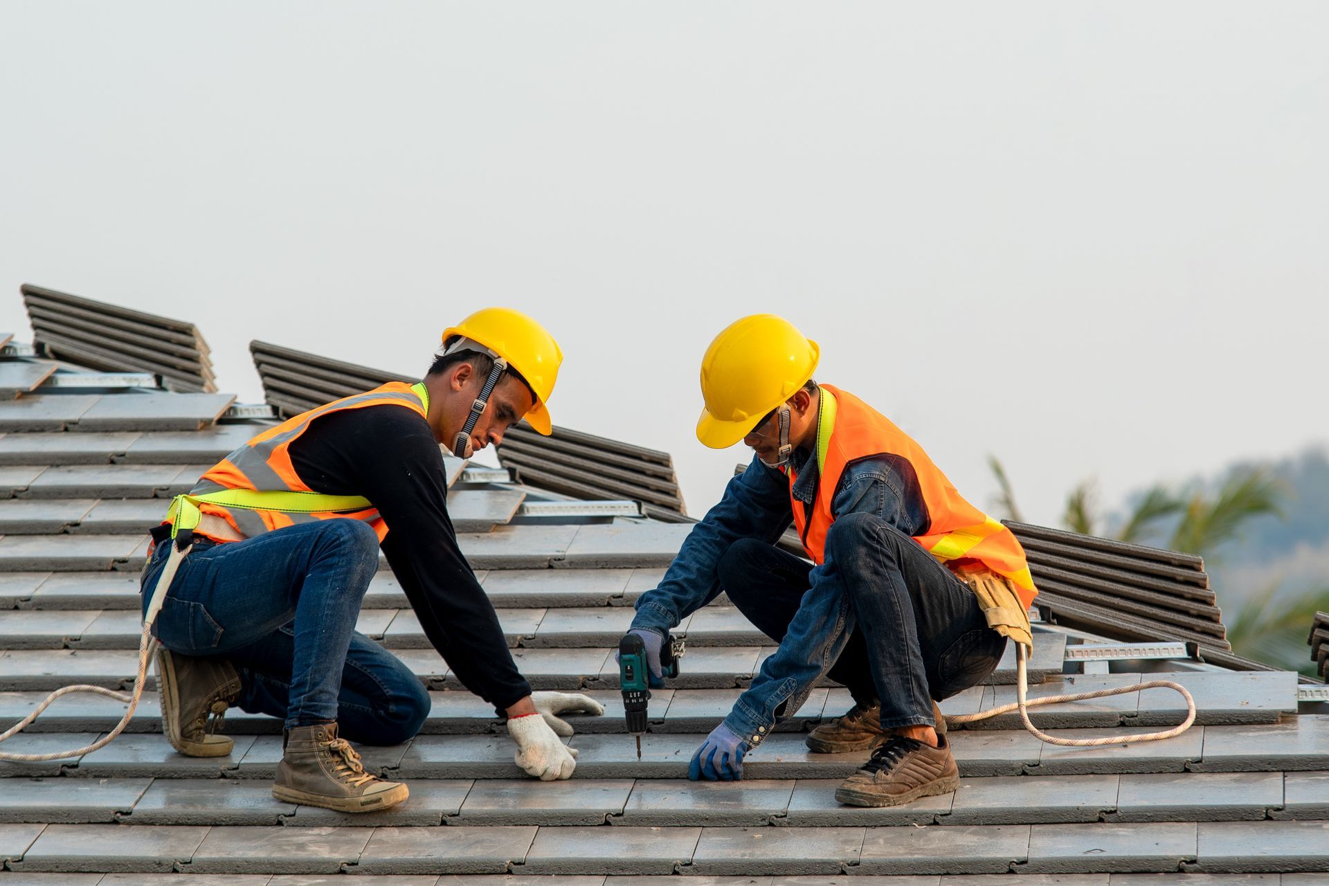 Two roofers in safety gear installing roof tiles with a power drill.