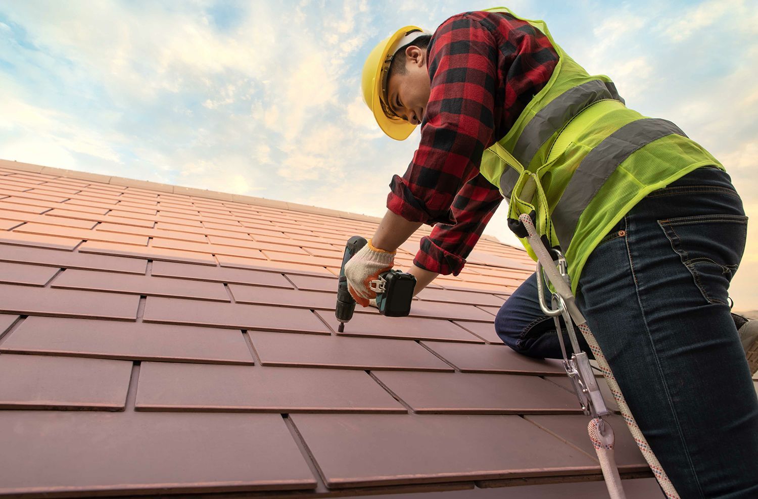 A residential roofing contractor installing shingles with a power drill on a house roof.