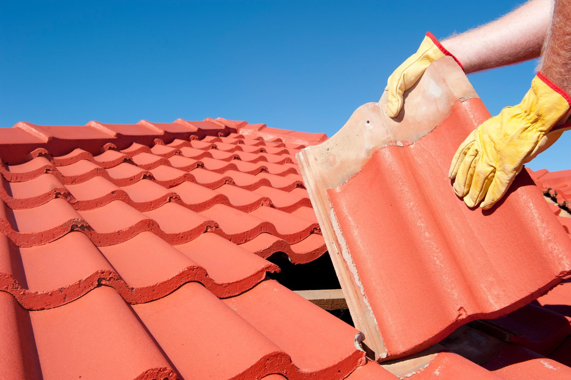 Gloved hands installing a red roof tile on a tiled roof under a blue sky