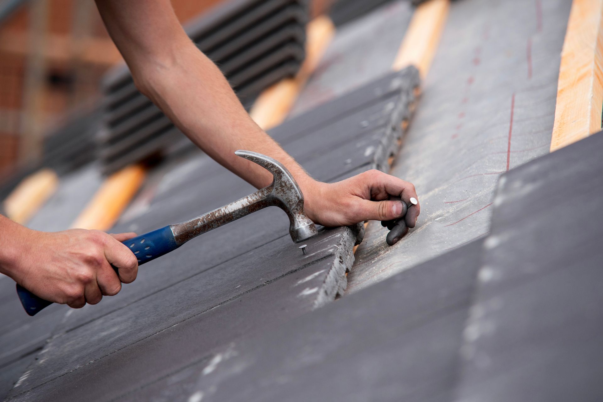 Hands nailing dark gray roofing shingles with a hammer on a rooftop