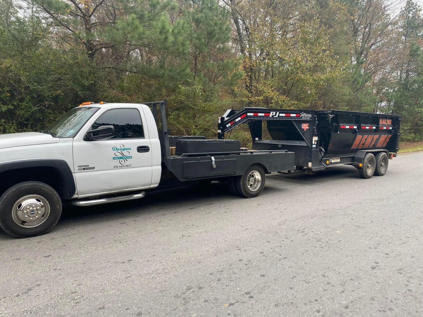 White pickup truck towing a black dump trailer on a gravel road beside trees