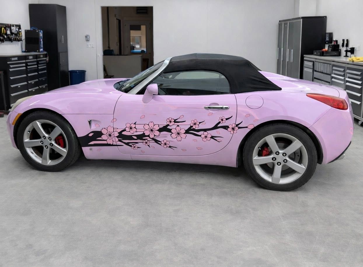 Pink convertible sports car with black roof and floral decals, parked in a garage.