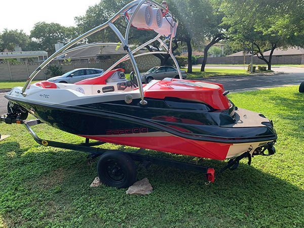 Red and black wakeboard boat on a trailer parked on grass in a sunny yard