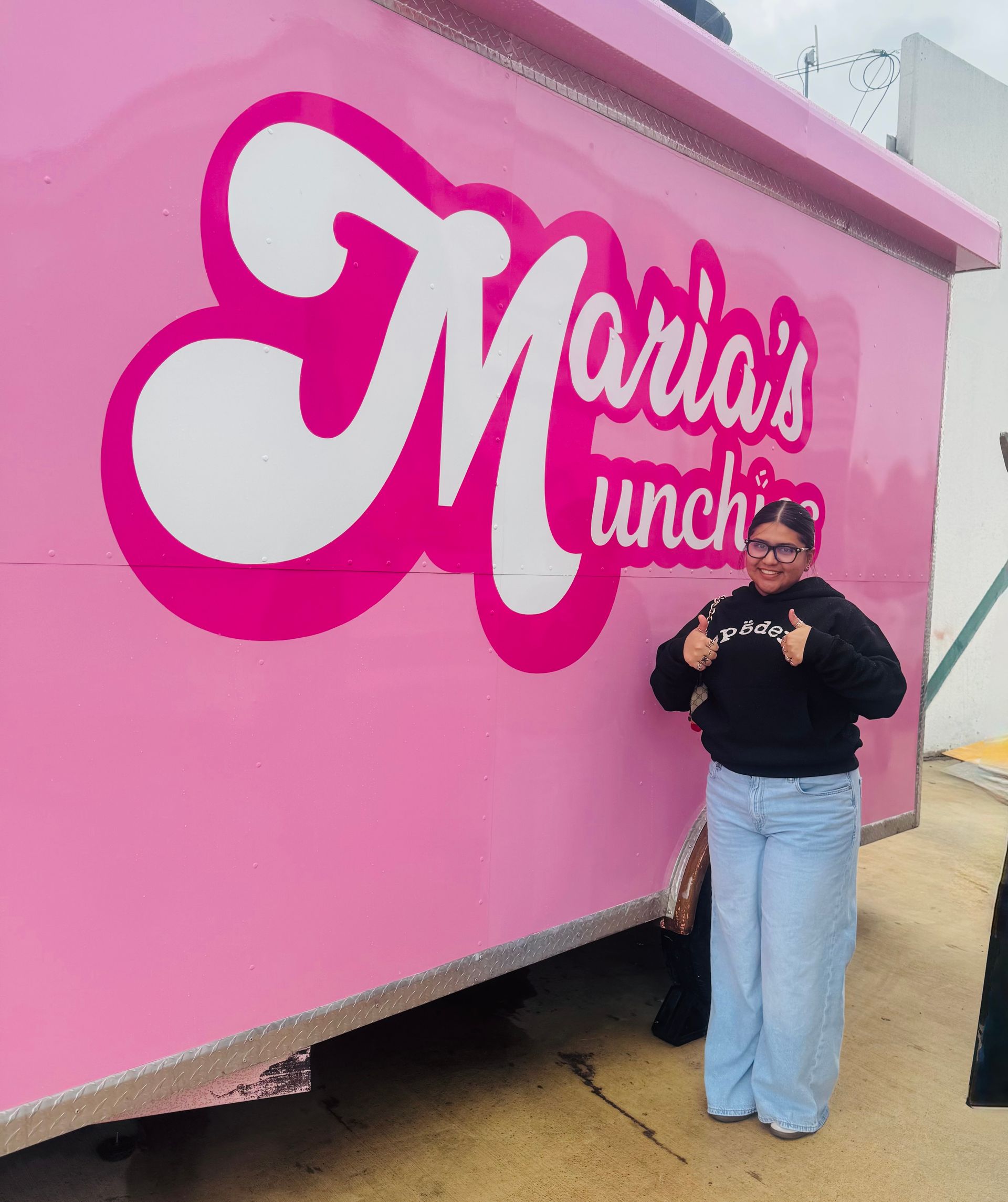 Woman standing beside a pink Maria's Lunch food truck, smiling and holding a drink.