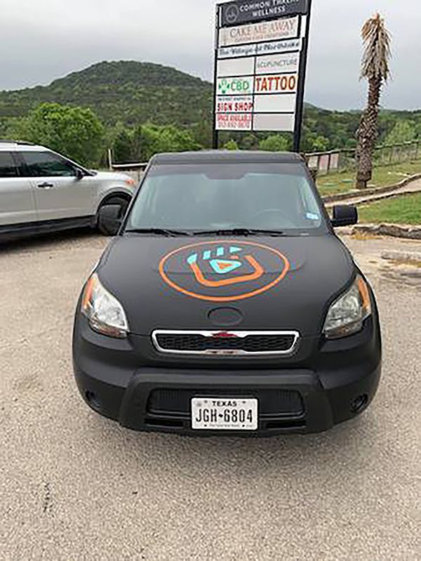 Black car with a colorful hood logo parked on a road near green hills and a roadside sign.
