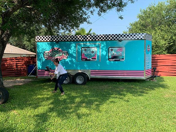 Turquoise food truck with pink stripes parked on grass; person walking in front under trees.