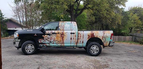 Rusty, weathered pickup truck parked on a gravel lot with trees and a fence in the background
