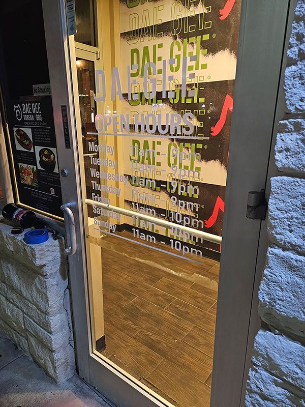 Glass storefront door with restaurant hours posted in white lettering and wooden floor visible inside.