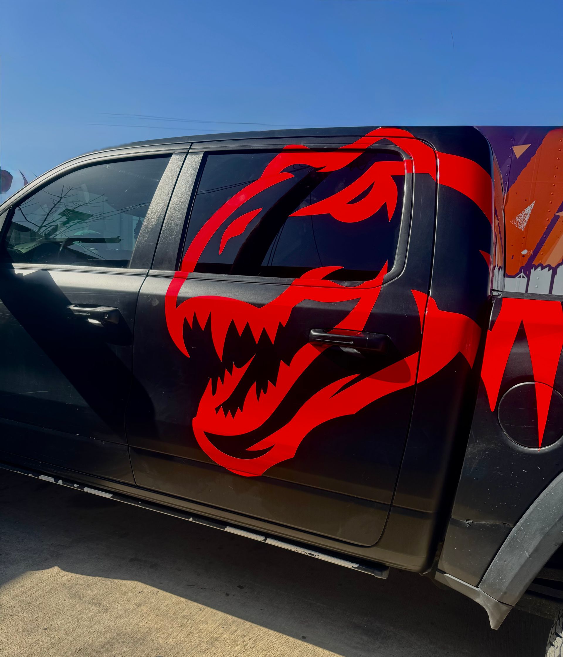 Black pickup truck with a large red dinosaur decal on the door, parked in sunlight.