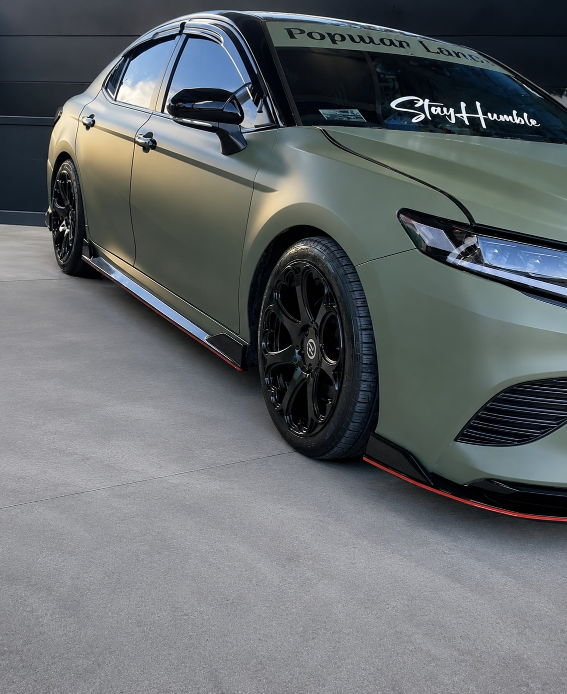 Matte olive sports sedan with black wheels and red accents parked outdoors.