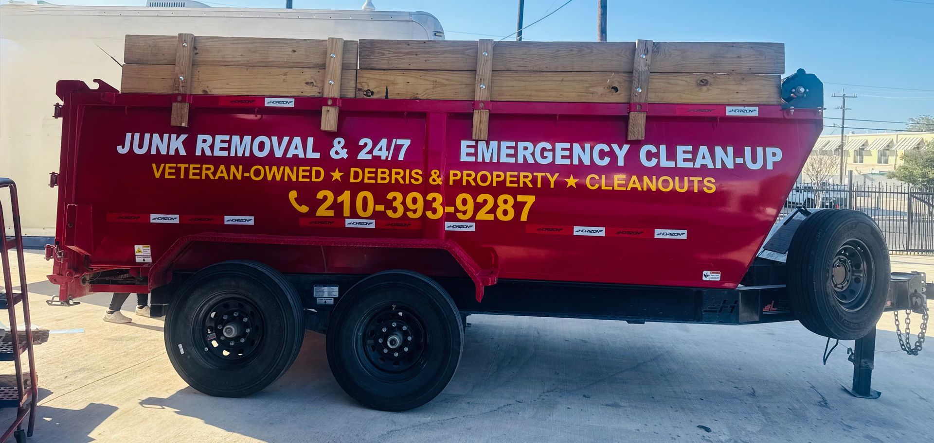 Red junk removal truck with trailer and contact number parked on a sunny street, hauling wooden debris.