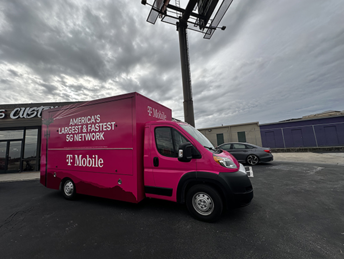 Pink T-Mobile van parked in a lot under cloudy skies, with a billboard overhead
