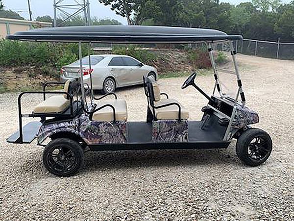 Black golf cart with camouflage seats and roof parked on gravel near a silver car