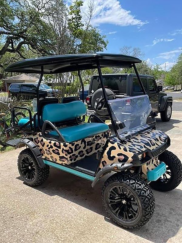 Leopard-print golf cart with teal seats parked outdoors on a sunny day.