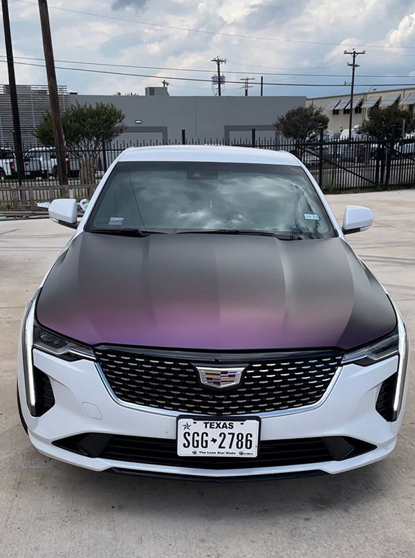 White Cadillac sedan with purple hood wrap, parked in front of a fence and industrial buildings.