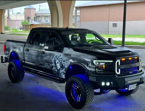 Black lifted pickup truck with blue underglow parked under a covered area, showing custom graphics and chrome accents