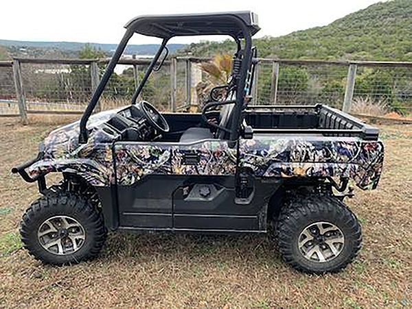Camouflage off-road utility vehicle parked on grass beside a fence and hills