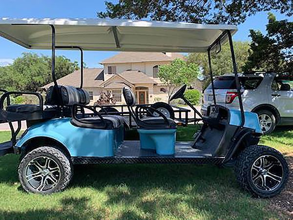 Blue golf cart parked in a driveway under a canopy, with a house and silver SUV in the background