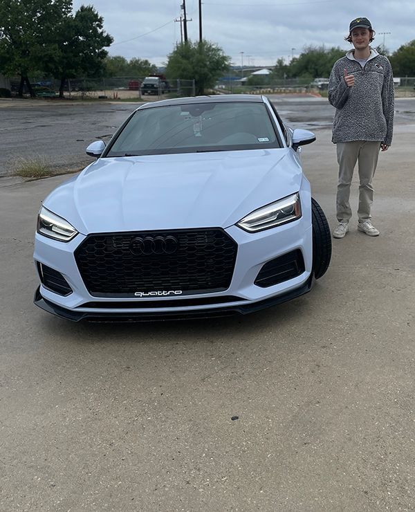 Person standing behind a light blue car in a parking lot on a cloudy day.