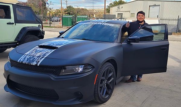 Person standing by an open door of a matte black Dodge Charger in a parking lot.