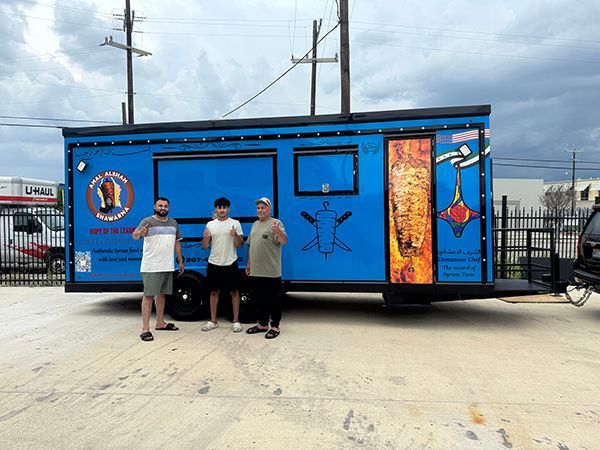 Three people stand in front of a bright blue food trailer with colorful graphics.
