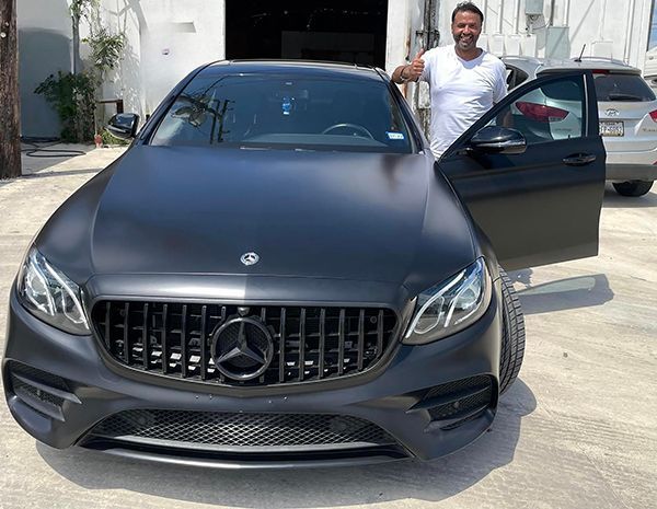 Matte black Mercedes-Benz parked outdoors with the driver door open and a man standing beside it
