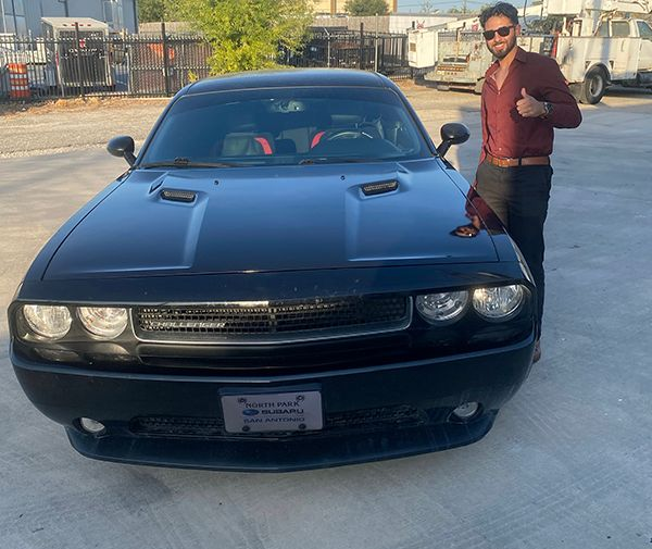 Man posing beside a black sports car in an outdoor lot, with sunlight and buildings in the background