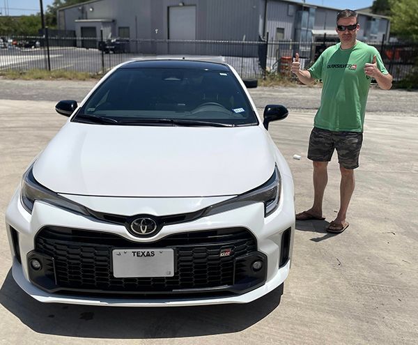 Man standing beside a white Toyota sedan in a sunny parking lot, making a thumbs-up gesture.