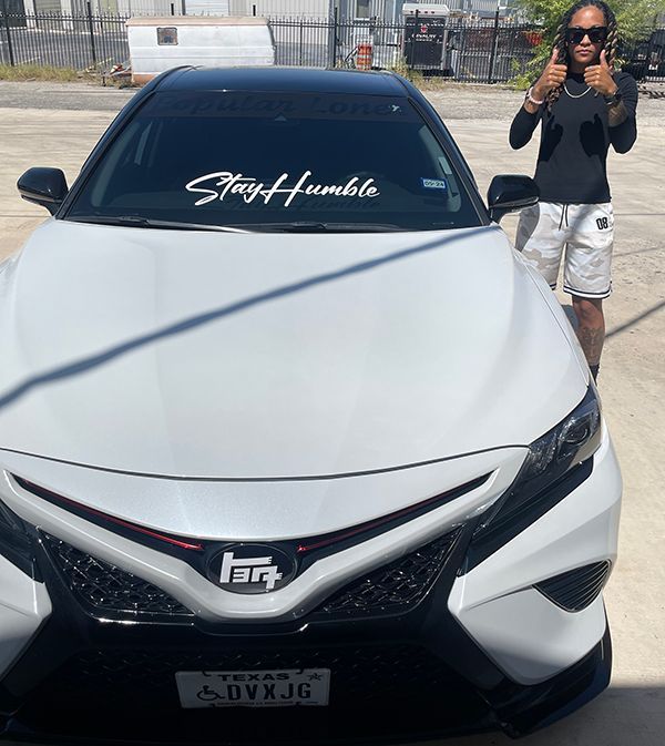 White sports car with tinted windows and custom windshield decal, parked beside a person posing outdoors.