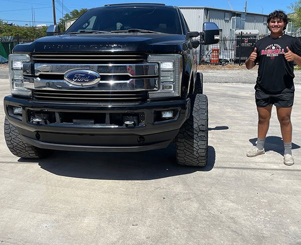 Person standing beside a black Ford truck in a sunny parking lot, giving a thumbs up.