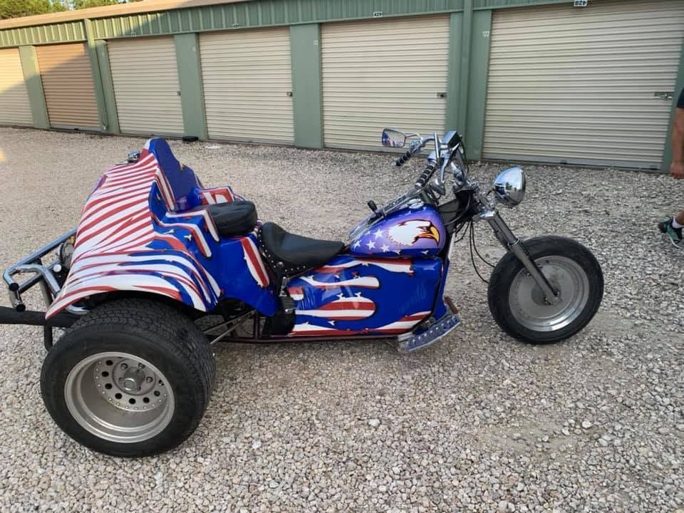 Blue three-wheeled motorcycle with red and white striped cover, parked on gravel by garage doors