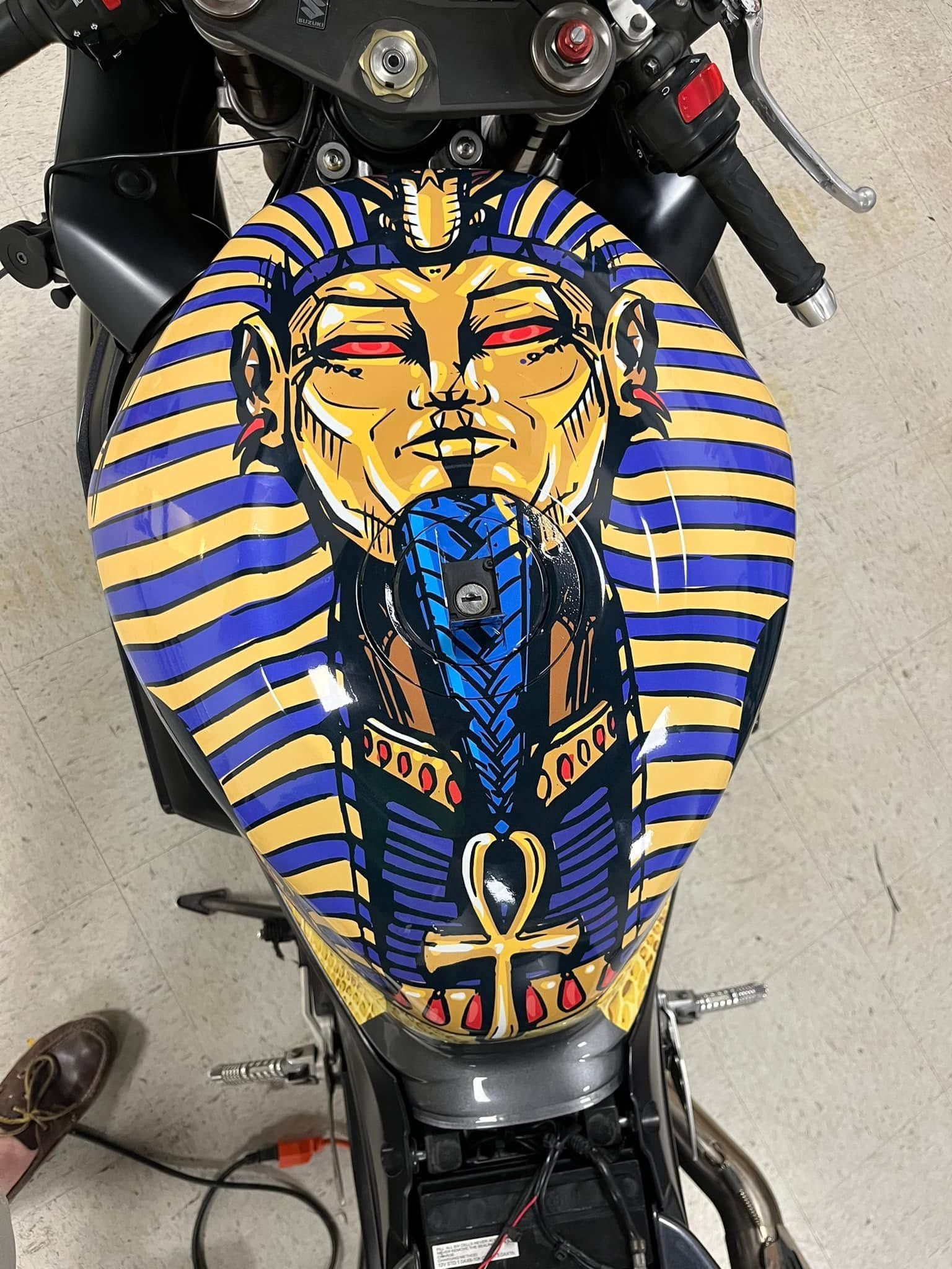 Motorcycle seat with a colorful pharaoh face design in blue, gold, and black stripes