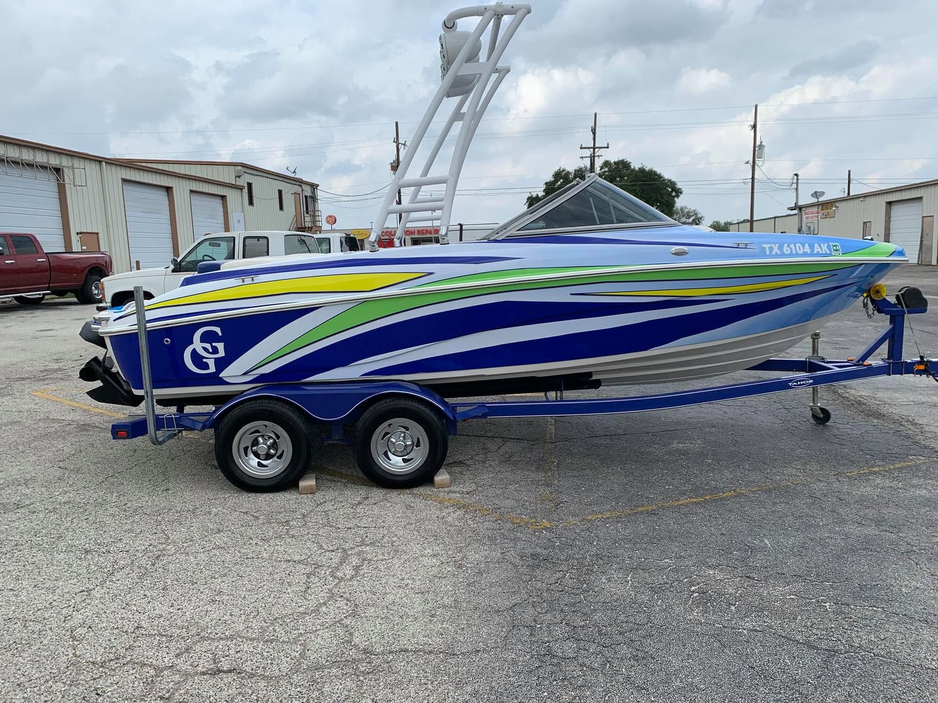 Blue and green speedboat on a trailer in a gravel lot, with industrial buildings in the background