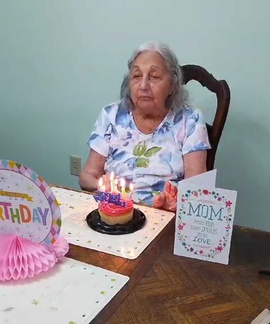 Woman seated at a table with a cupcake and birthday decorations. She looks at the lit candles.