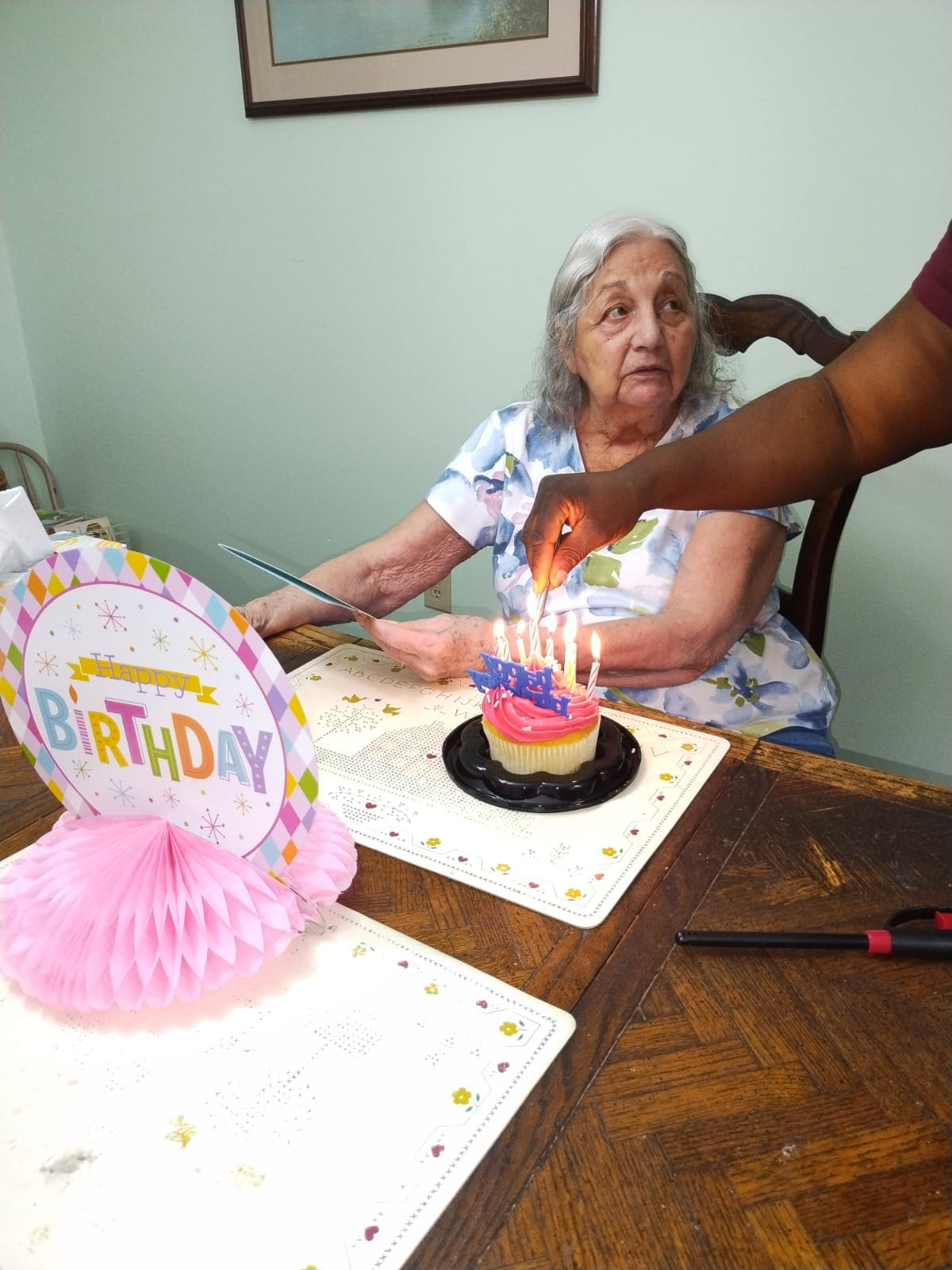 An older person sits as a hand lights candles on a cupcake. A birthday sign sits on the table.