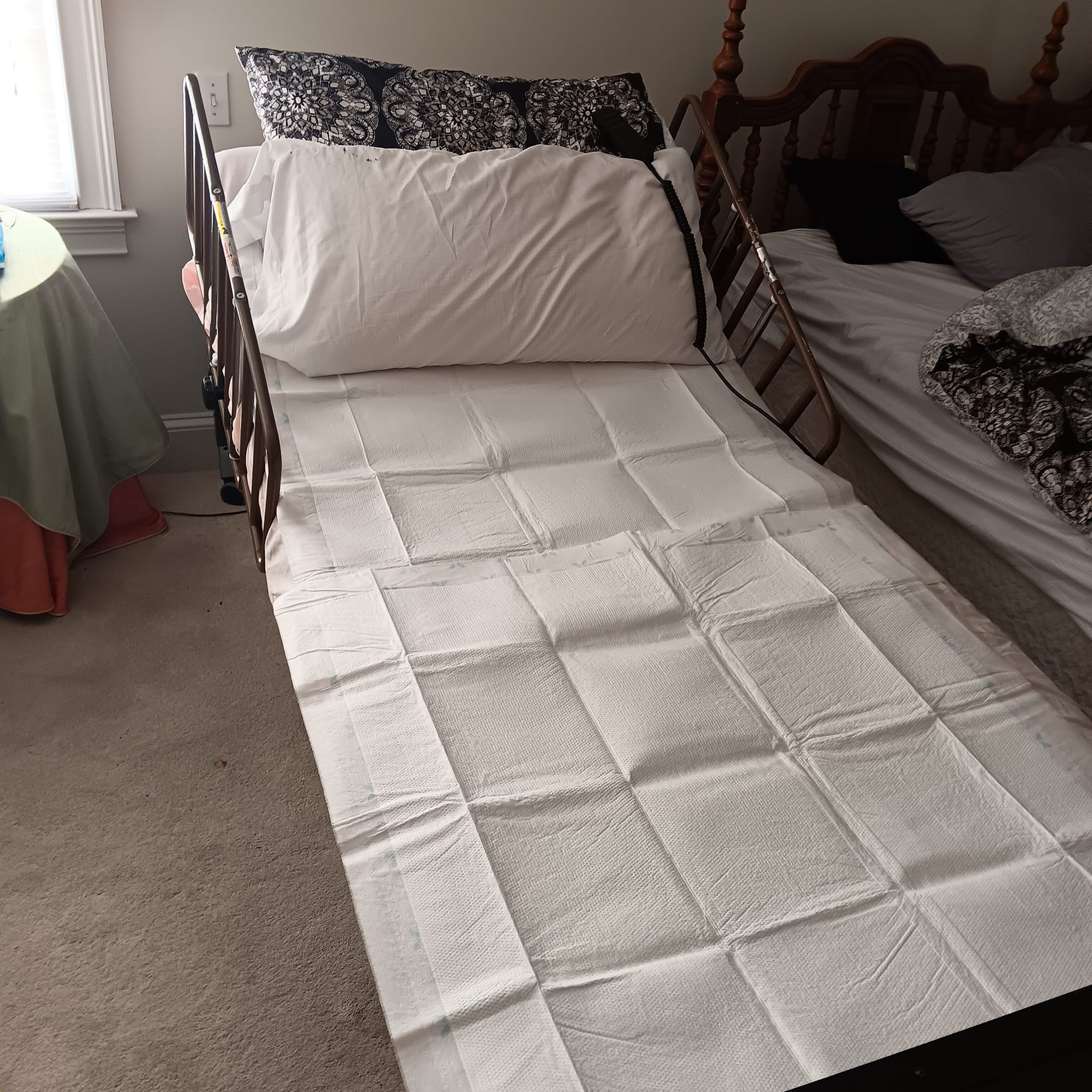 A hospital bed with white quilted pads, pillows, and a nearby table covered with a green cloth.
