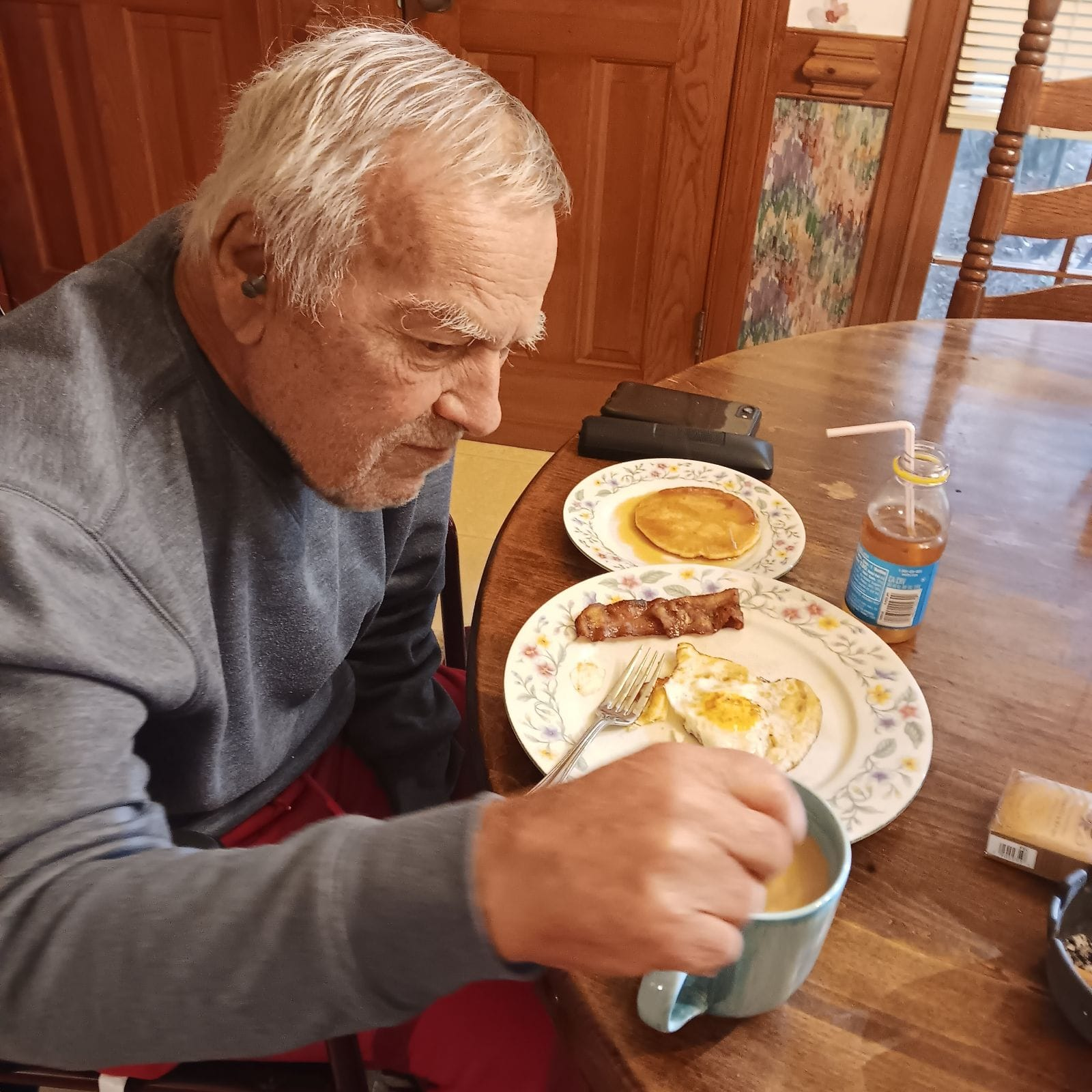 Man seated at a table eating breakfast: eggs, sausage, pancake, coffee, apple juice.
