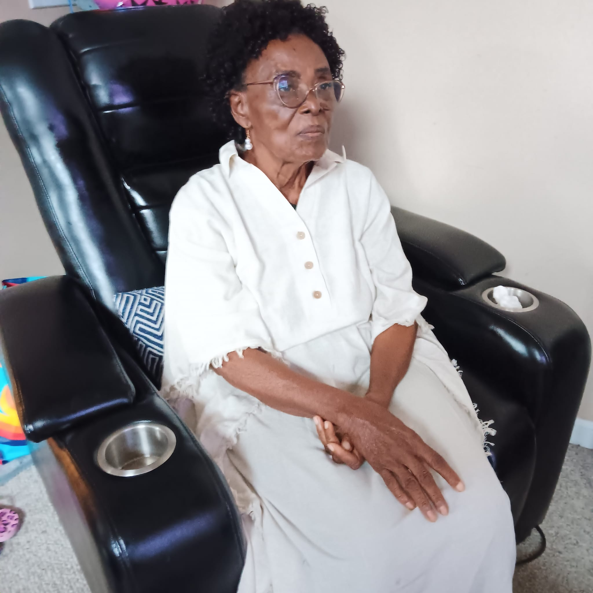 Woman in glasses sits in a black recliner chair, hands clasped, light-colored clothing.