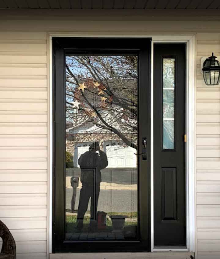 Front door with tall glass panel reflecting a person and trees; beige siding and black trim.