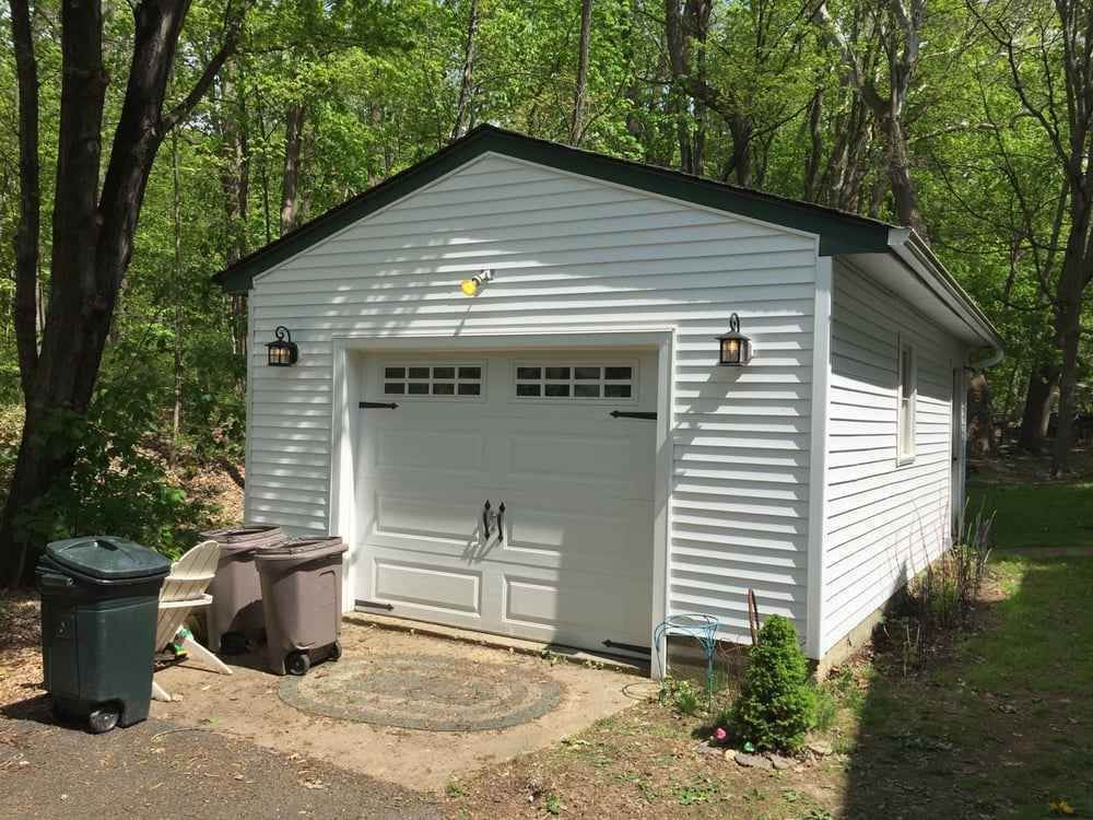 White detached garage with a closed door in a wooded backyard, with trash bins beside it.