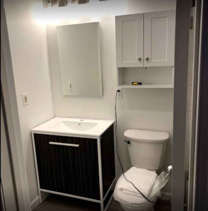 Small bathroom vanity and toilet with white walls, gray cabinets, and a cluttered laundry bag on the floor.