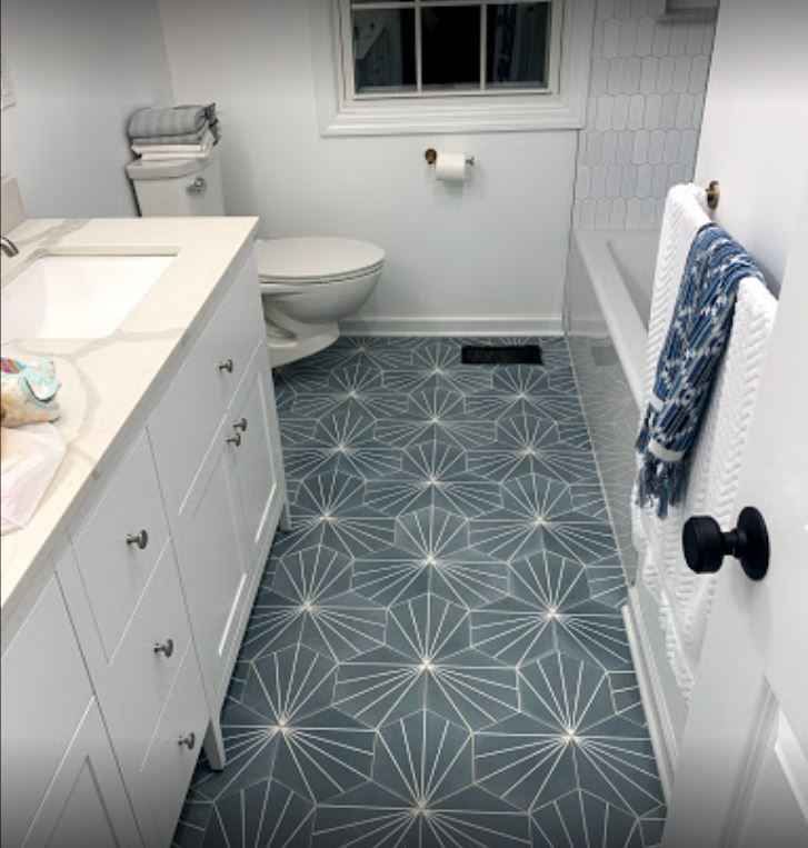 Small bathroom with blue patterned floor, white vanity, toilet, window, and towel rack.