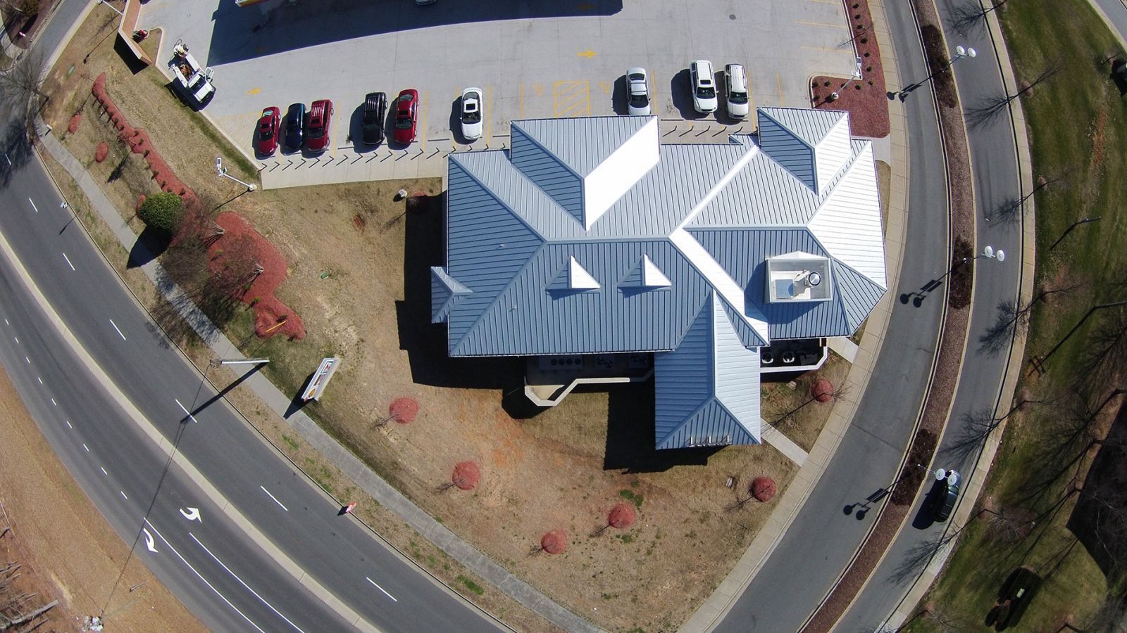 Aerial view of a building with a silver roof beside a curved road and parking lot with several cars.