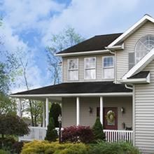 Two-story suburban house with white siding, dark roof, front porch, and manicured shrubs under a blue sky