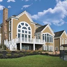Large tan suburban house with white trim, arched windows, and a front porch under a blue sky