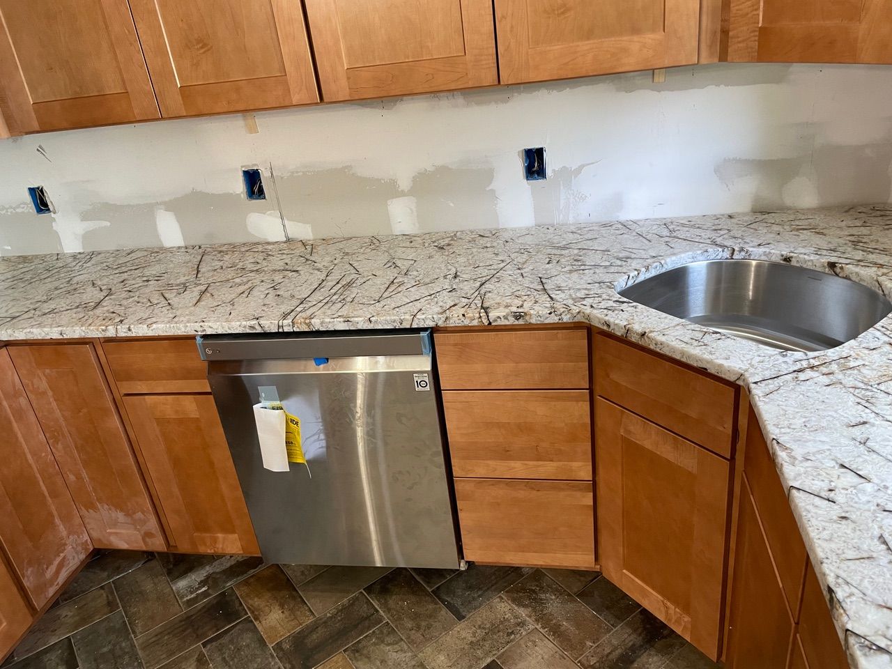 Kitchen corner with granite countertop, wooden cabinets, stainless dishwasher, and sink under unfinished backsplash area