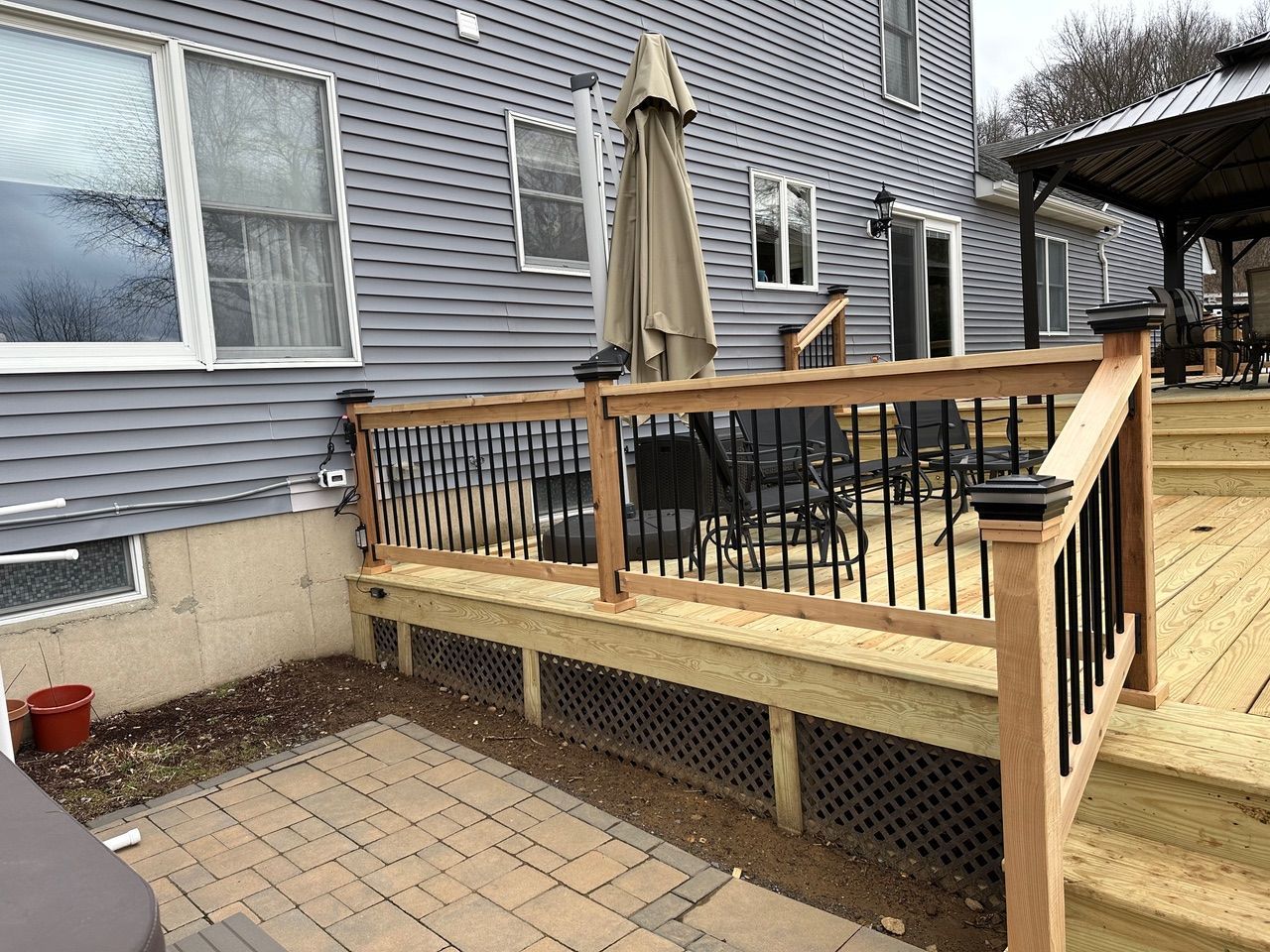 Backyard deck with wooden railing, patio pavers, and a closed umbrella beside a gray house