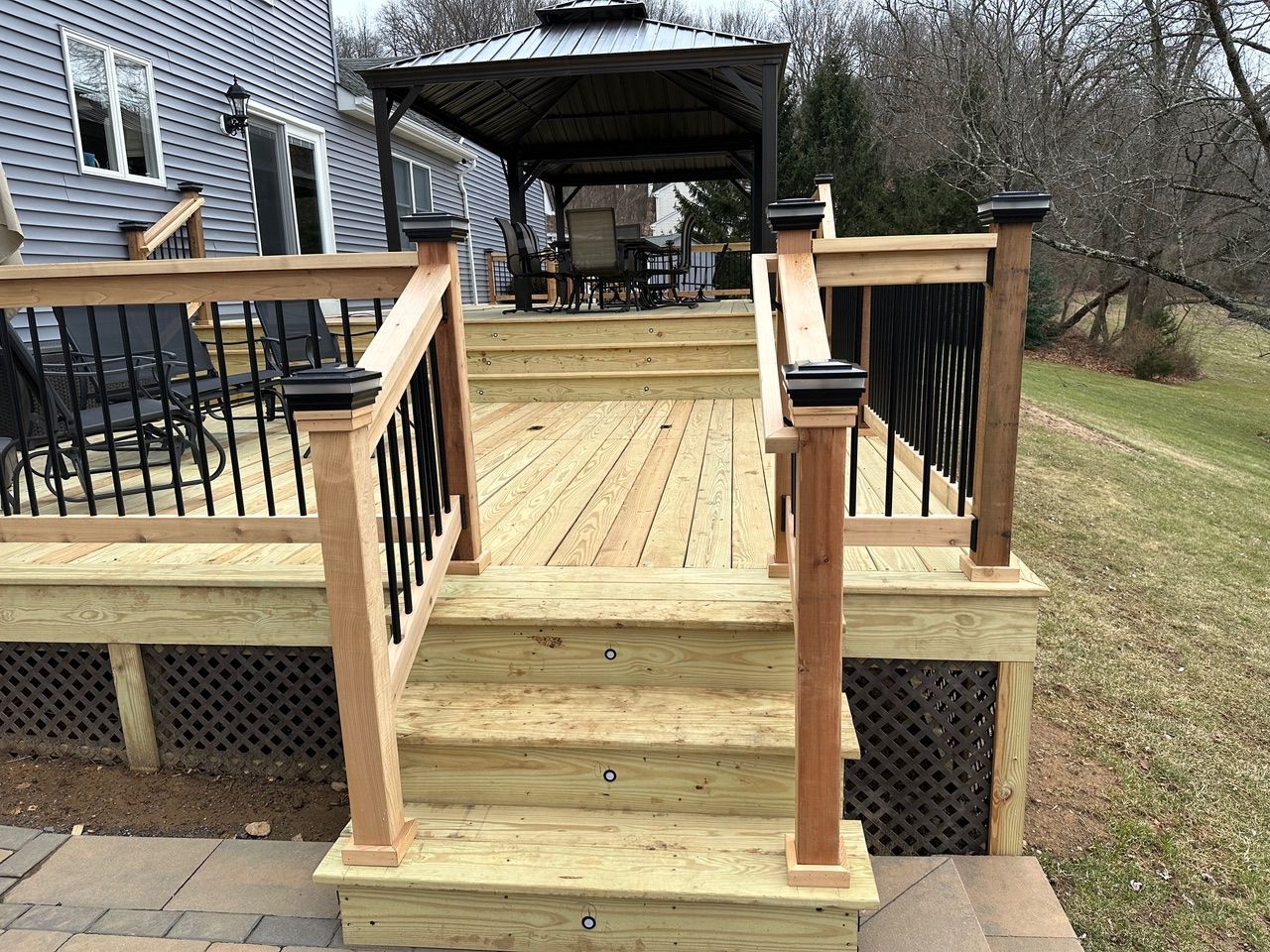 Wooden deck with steps and black railings beside a gray house, overlooking a grassy yard