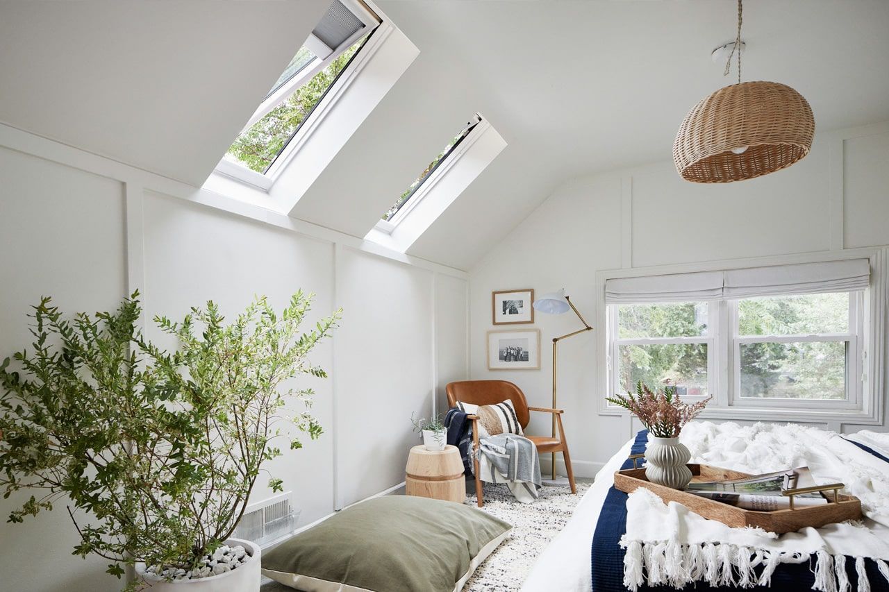 Bright, airy bedroom with skylights, white walls, plants, a woven pendant lamp, and a cozy bed by the window.